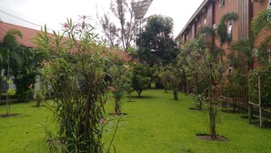 A courtyard with green grass, various plants, and a building with a red roof in the background. - Myanmar Sports Hotel (Yangon)