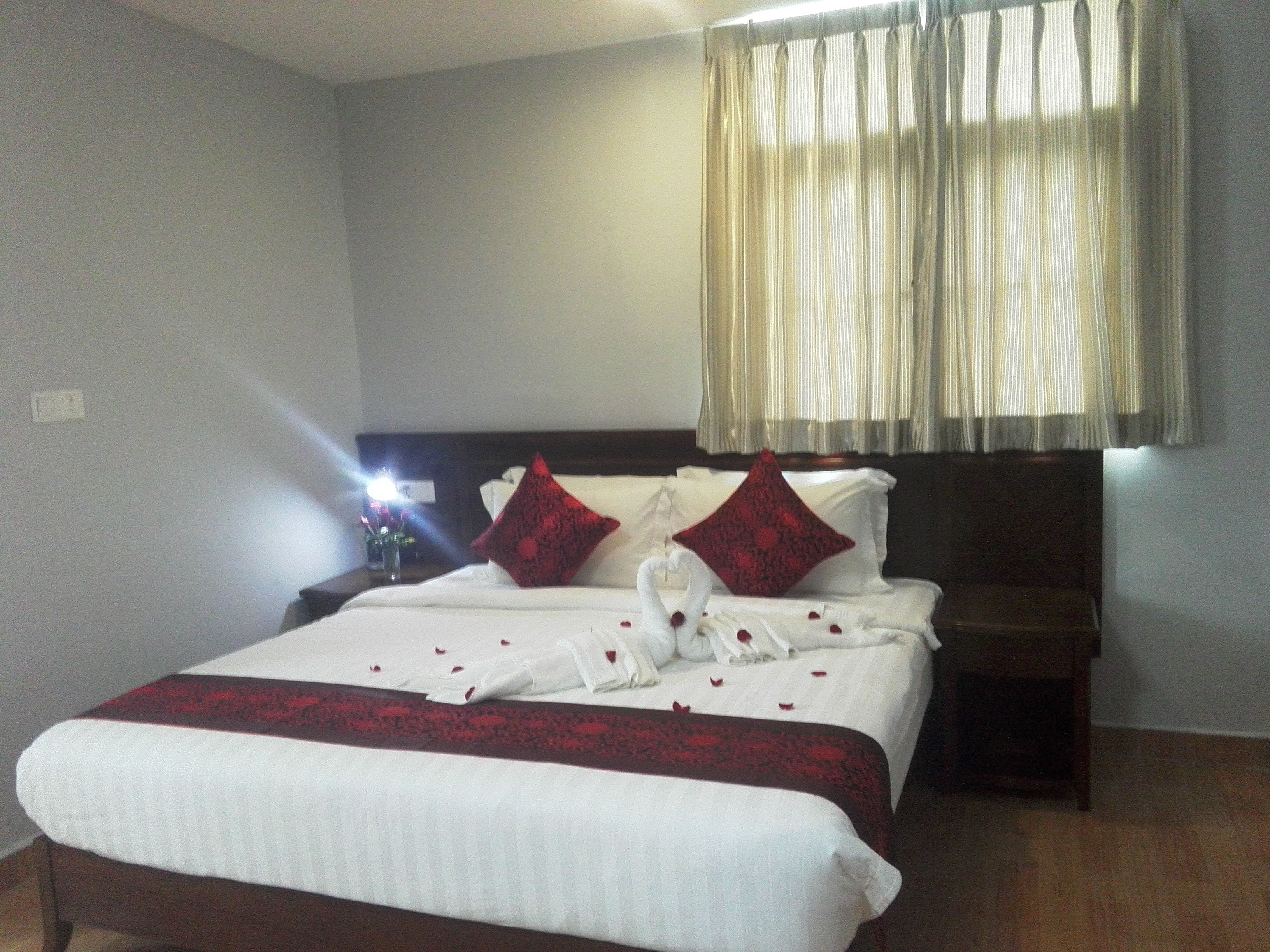 Habitación de hotel con una cama grande, una mesita de noche, un florero con flores y una ventana con cortinas.