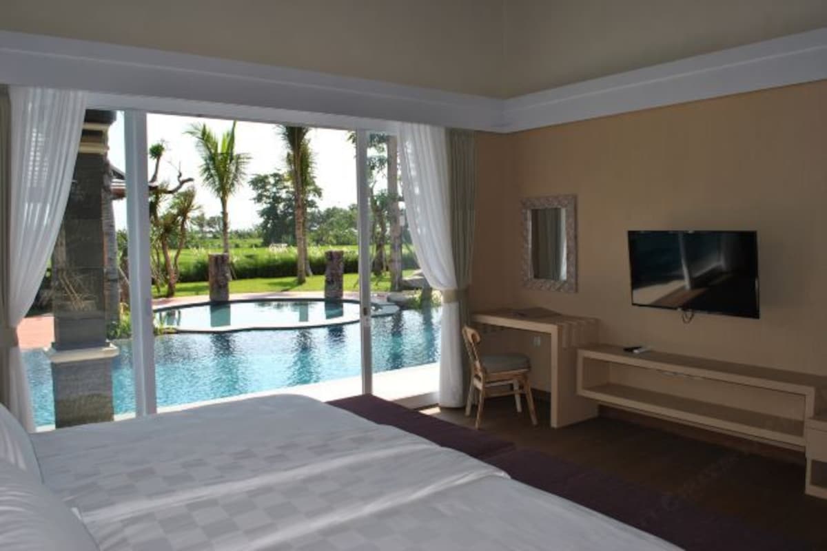 Hotelkamer met een groot bed, een bureau, een stoel, een televisie en uitzicht op een zwembad en groen.
