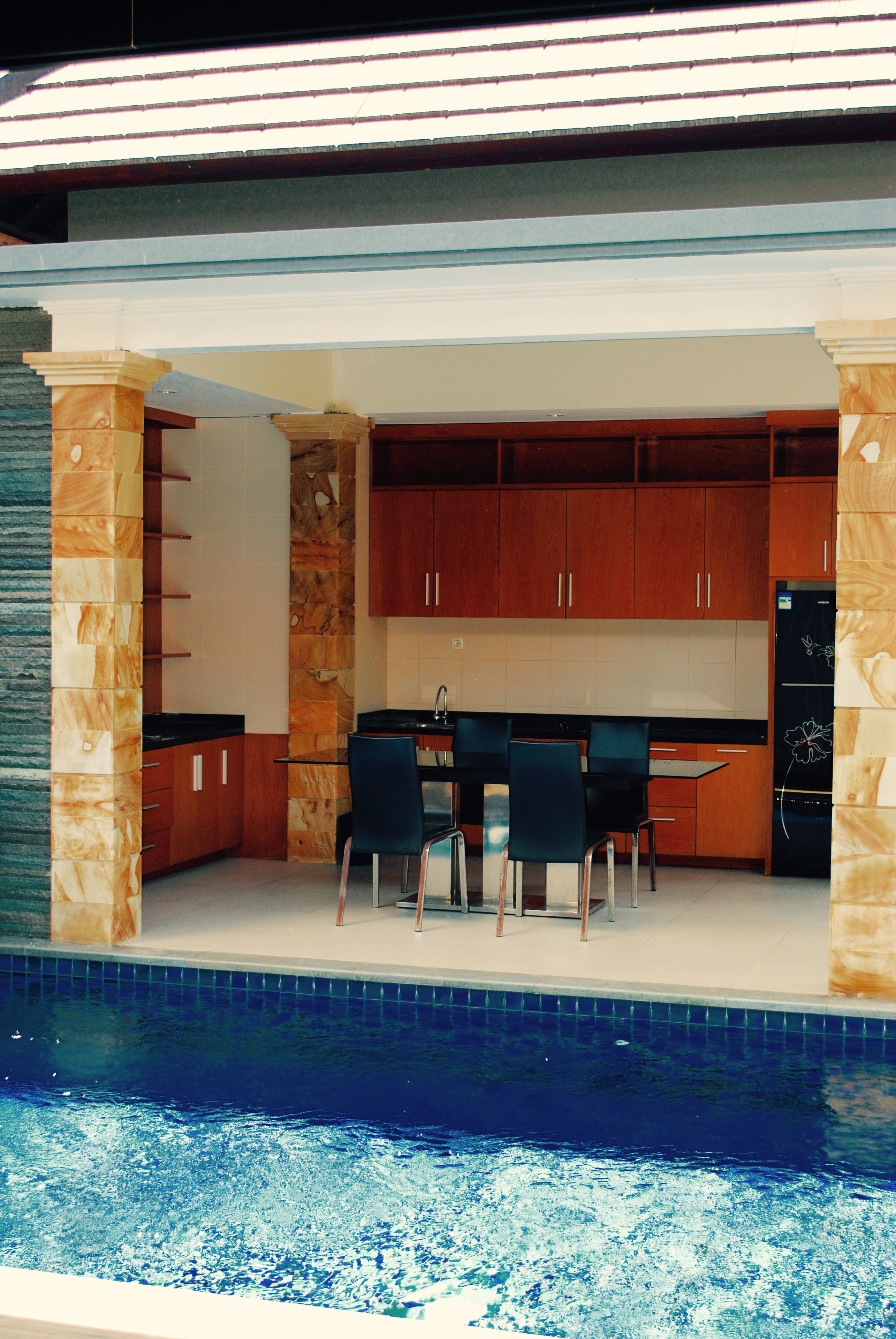 A modern kitchen with wooden cabinets and a built-in refrigerator, adjacent to a swimming pool with a dining area.