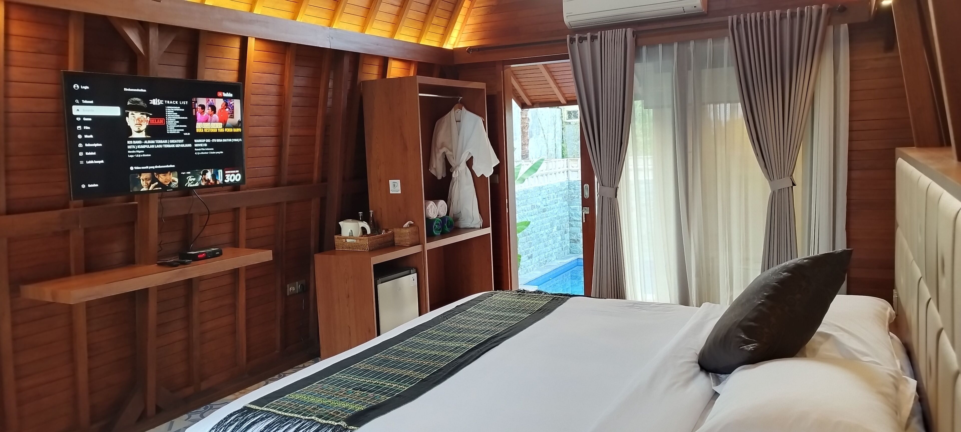 A wooden room with a bed, a TV displaying a track list, and a view of the outside.