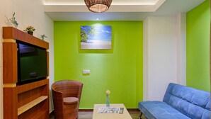 Una sala de estar con televisor, una silla de mimbre, un sofá azul y una pared verde con una pintura de playa.