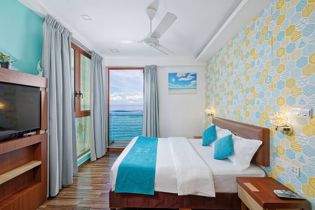 A hotel room with a bed, a TV, a ceiling fan, and a view of the sea.