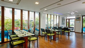 Un área de comedor moderna con piso de madera, sillas acolchadas color verde y amplias ventanas que ofrecen vistas a la vegetación y edificios.