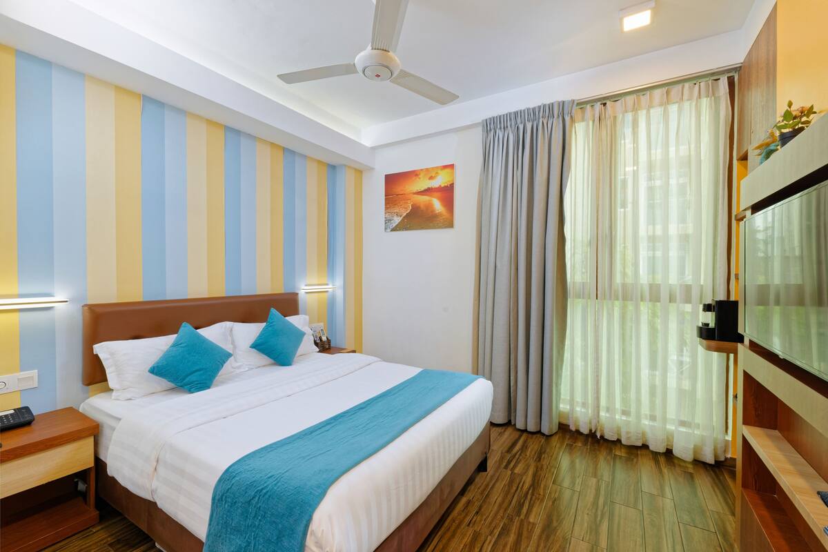 A hotel room with a bed, a ceiling fan, a painting on the wall, and a TV on the wall.