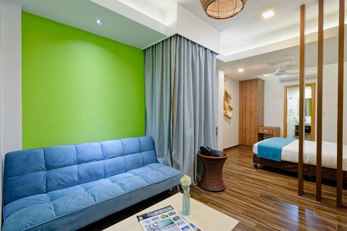 A modern hotel room with a blue sofa, a bed with a blue blanket, a wicker side table, and a green accent wall.