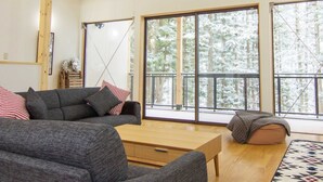 A modern living room with a sofa, a coffee table, and a view of a snowy forest through large windows. - Morino Lodge Chalets (Hakuba)