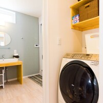 Una sala de lavandería con lavadora, un estante de madera con una cesta, y un baño con lavamanos y espejo.