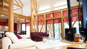A modern living room with a white sofa, a red sofa, and a glass coffee table. There is a fireplace with a black chimney and a wooden ceiling structure. - Morino Lodge Chalets (Hakuba)