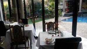 Un espace de restauration en plein air avec des tables pour deux, donnant sur une piscine et un jardin.