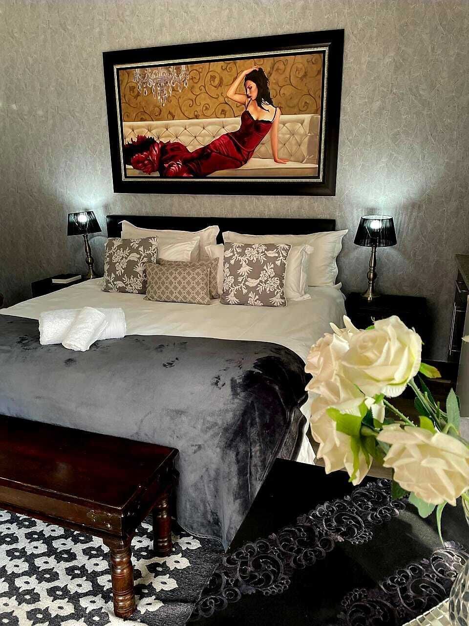 Un dormitorio con cama, almohadas, una mesita de noche, una lámpara, un florero con flores y un cuadro en la pared.