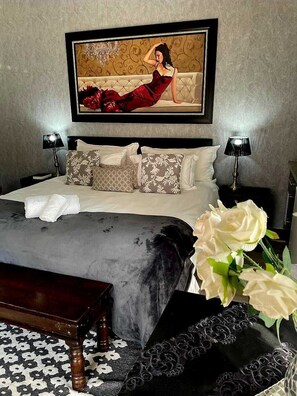 A bedroom with a bed, pillows, a bedside table, a lamp, a vase with flowers, and a painting on the wall.