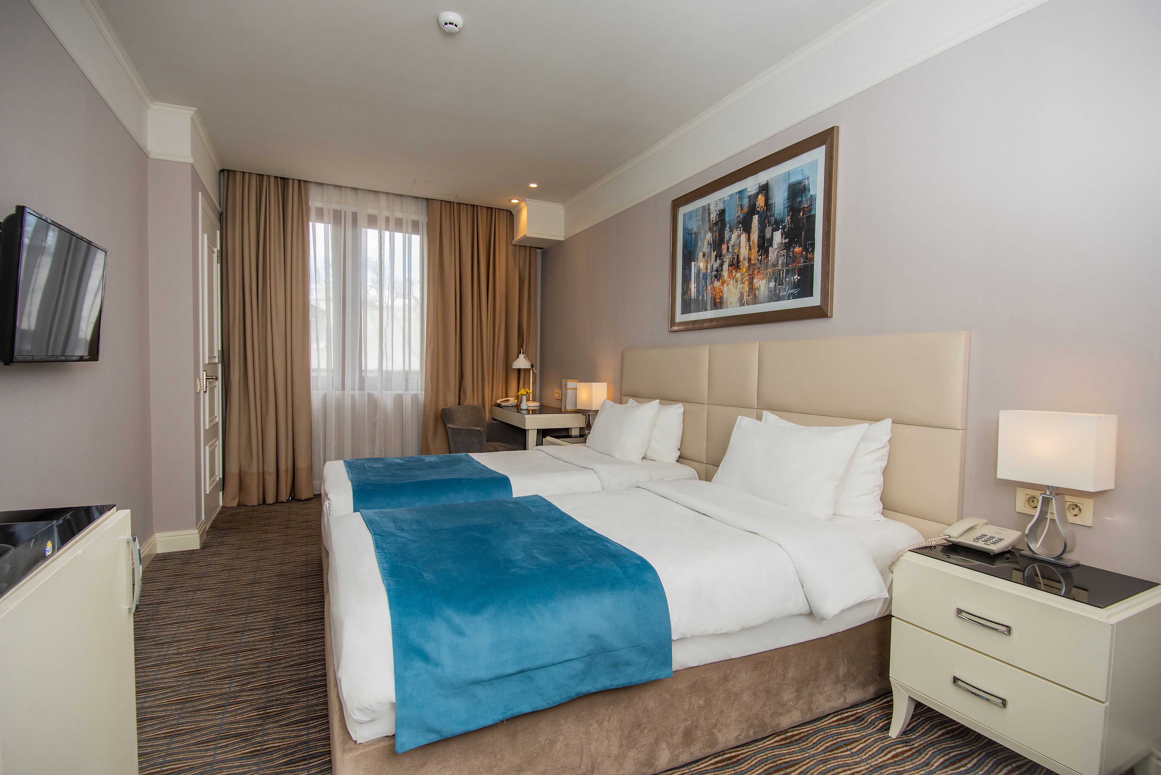 Quarto de hotel com uma cama grande, uma mesa com luminária, uma televisão e um quadro na parede.