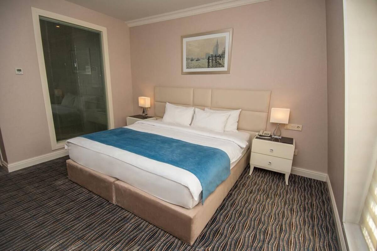 Ein Hotelzimmer mit einem großen Bett, einem Nachttisch mit Lampe, einem Wandbild und einem gemusterten Teppich.