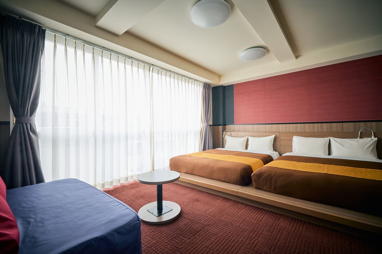 Hotellihuone, jossa on kaksi sänkyä, pyöreä pöytä ja suuri ikkuna, jossa on verhot.