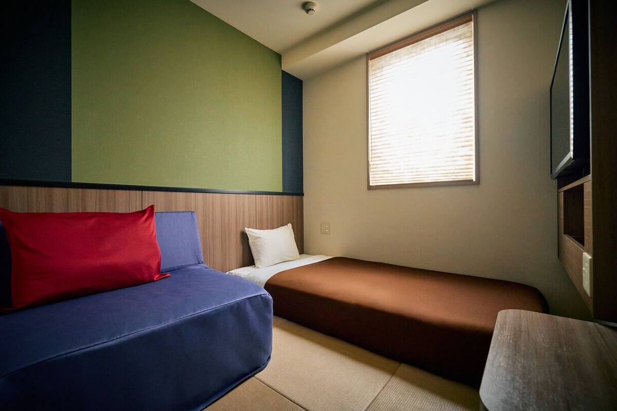 A room with a bed, a sofa, a window with blinds, and a television mounted on the wall.