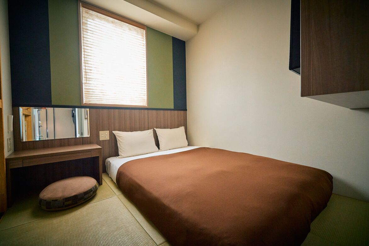 A small, neatly arranged bedroom with a single bed, a bedside table, a chair, and a window with blinds.