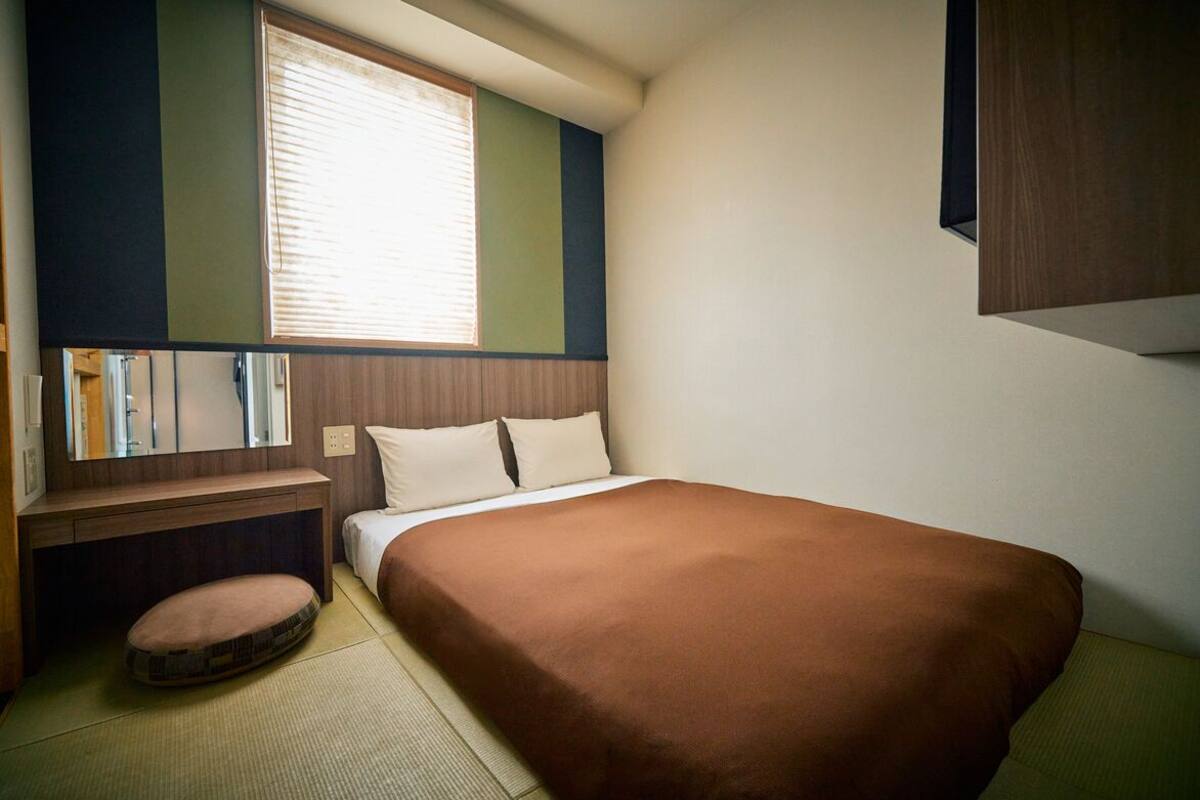 A small, neatly arranged bedroom with a single bed, a bedside table, a chair, and a window with blinds.