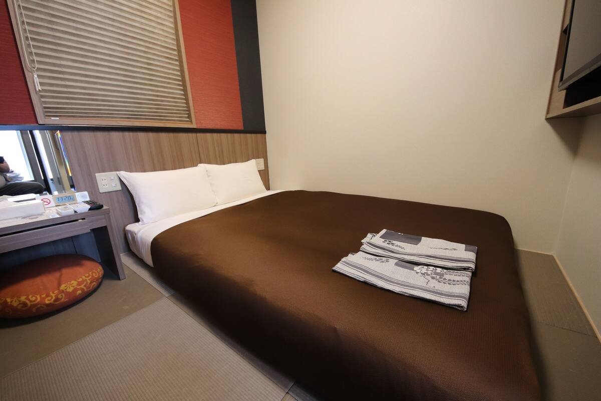 A hotel room with a bed, a desk with a chair, a window with blinds, and a wall-mounted TV.