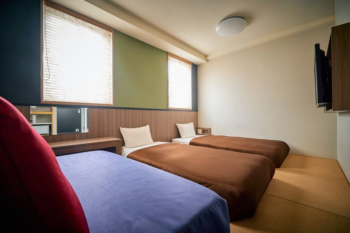 A hotel room with two beds, a TV, a window with blinds, and a ceiling light.