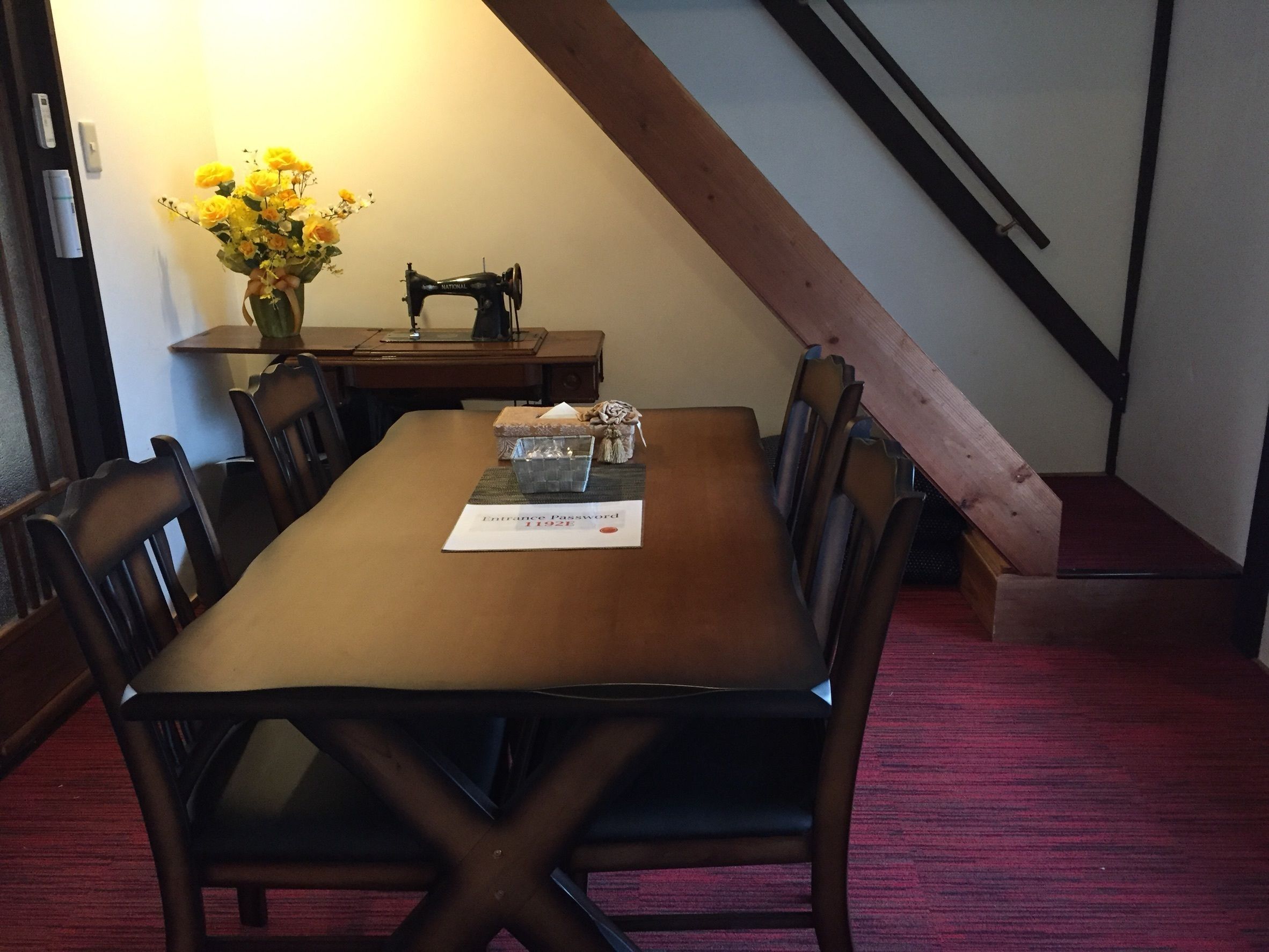 A dining area with a wooden table, chairs, a vase of flowers, a sewing machine, and a red carpet.