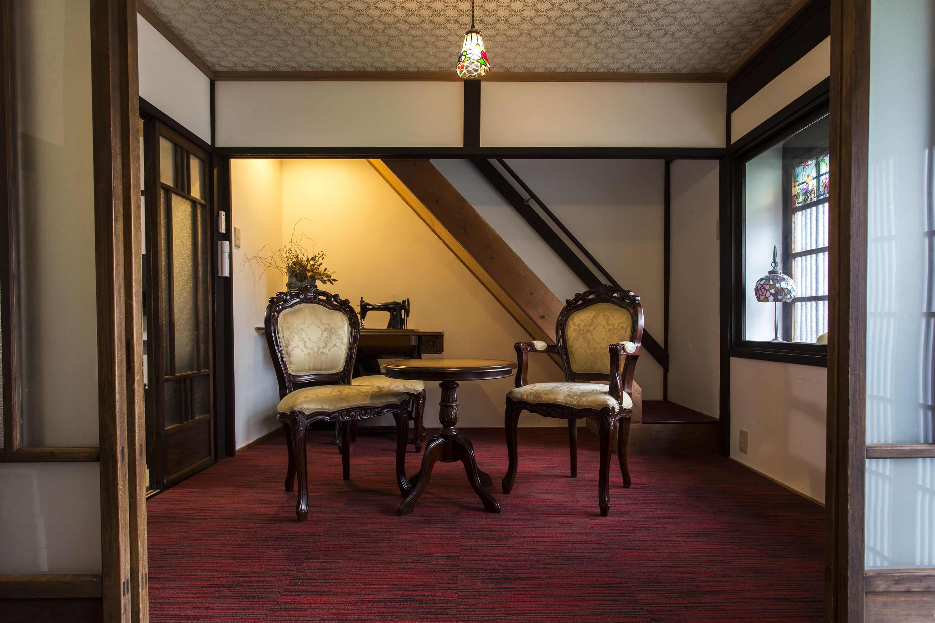 A room with a red carpet, two chairs, a small table, and a staircase.