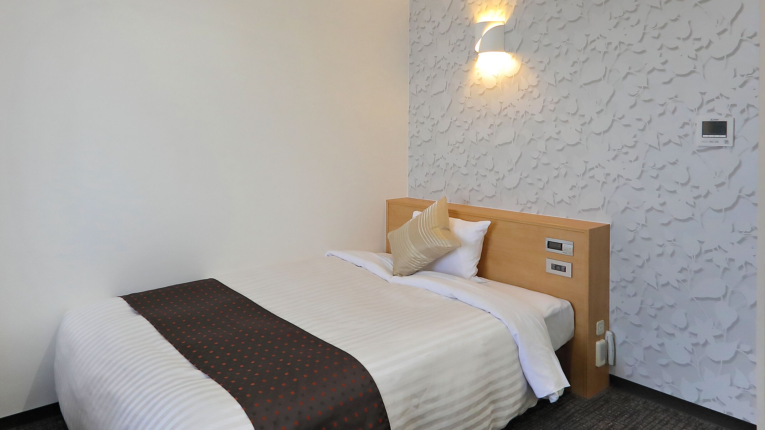 A hotel room with a bed, a headboard with controls, a wall-mounted light, and a thermostat.