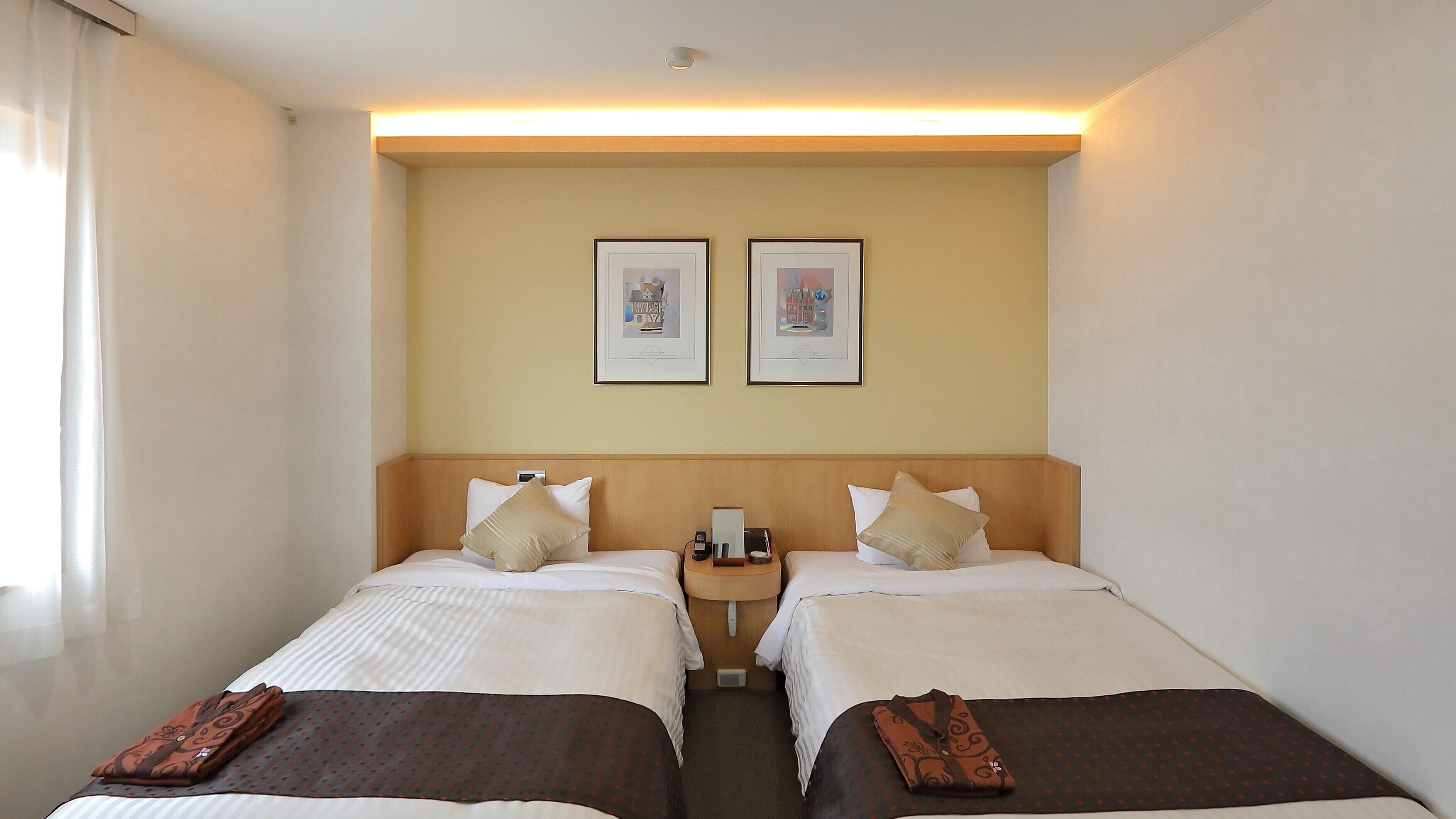 A hotel room with two single beds, a bedside table with a lamp, a wall-mounted phone, and two framed pictures above the beds.