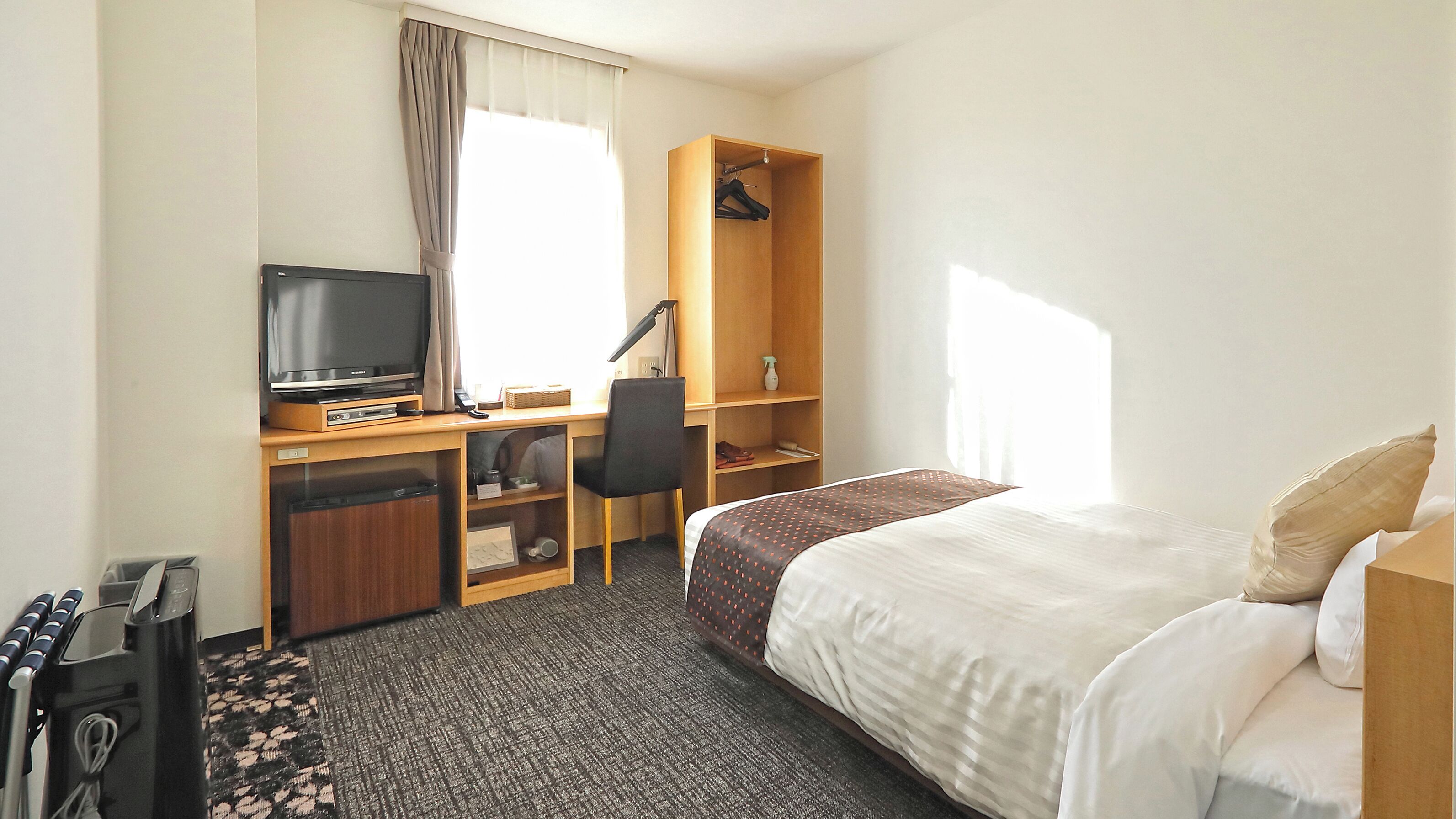 A hotel room with a bed, a desk with a television, a chair, a wardrobe, and a carpeted floor.