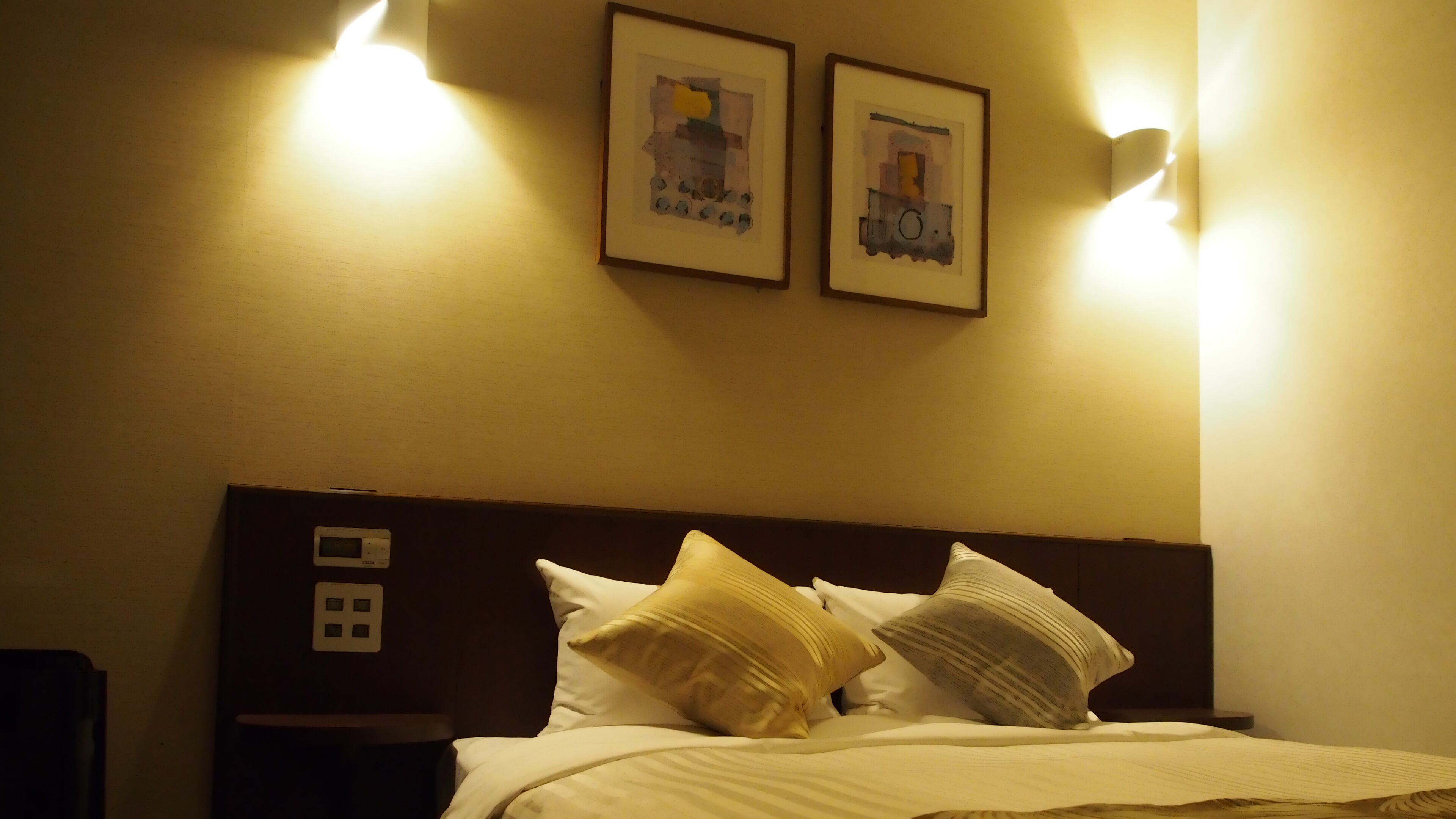 A hotel room with a bed, two wall-mounted light fixtures, and two framed pictures on the wall.