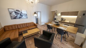 A modern living room with a brown leather sofa, a wooden coffee table, and black chairs. A dining area with a wooden table and chairs is adjacent to the living room. A kitchen with a refrigerator, microwave, and sink is visible. A chandelier hangs from the ceiling, and a framed picture is on the wall. - New Town - Apple Apartments (Prague)