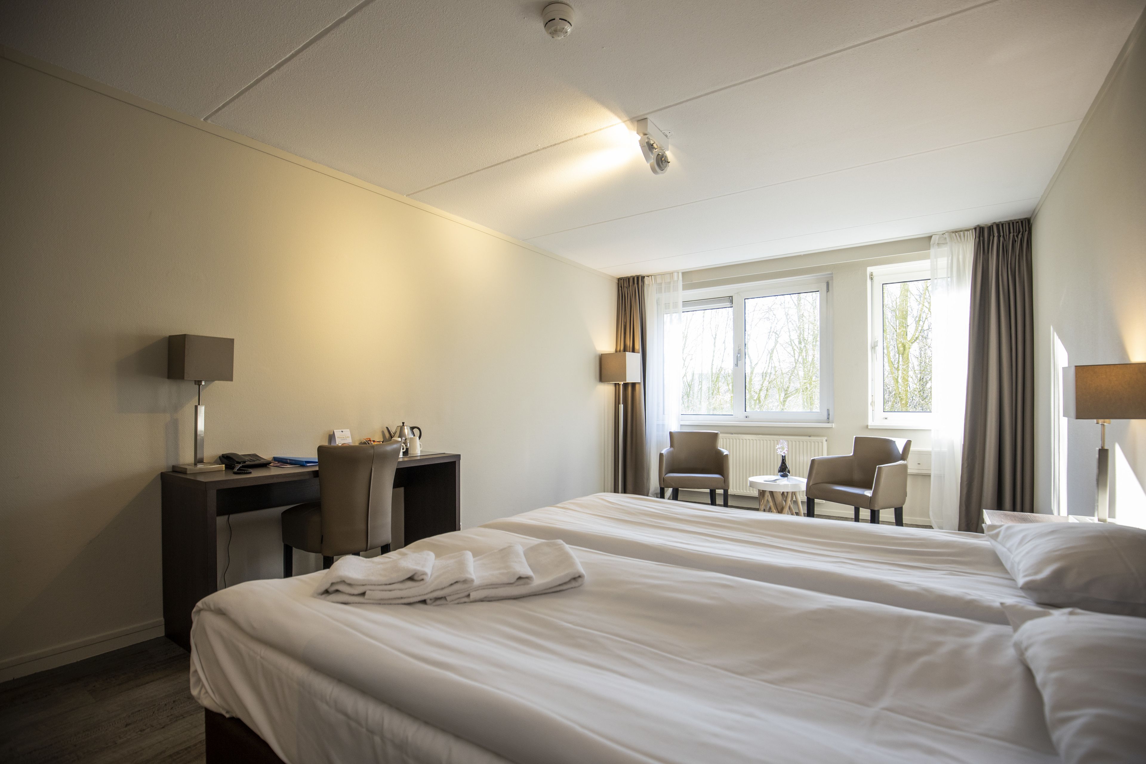 A hotel room with two beds, a desk, and a chair. There is a window with curtains, a lamp, and a small table with a chair.