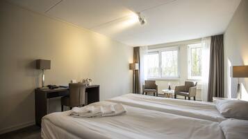 Een hotelkamer met twee bedden, een bureau en een stoel. Er is een raam met gordijnen, een lamp en een klein tafeltje met een stoel.
