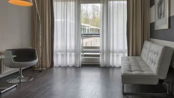 A modern hotel room with a sofa, a chair, a floor lamp, and a large window with curtains.