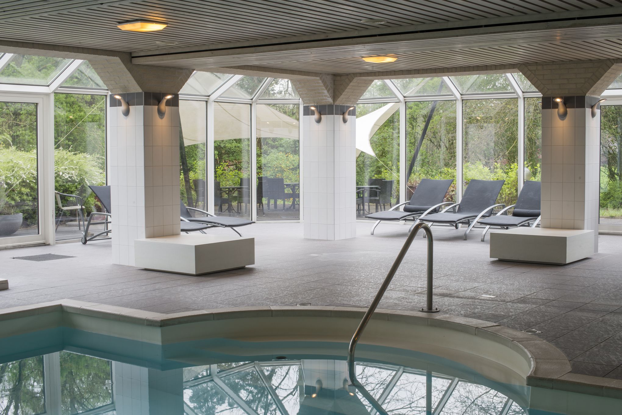 A modern indoor pool area with clear glass walls, lounge chairs, and a central pool.