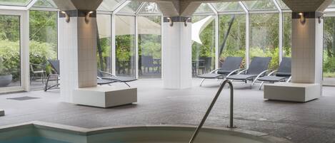 A modern indoor pool area with clear glass walls, lounge chairs, and a central pool.