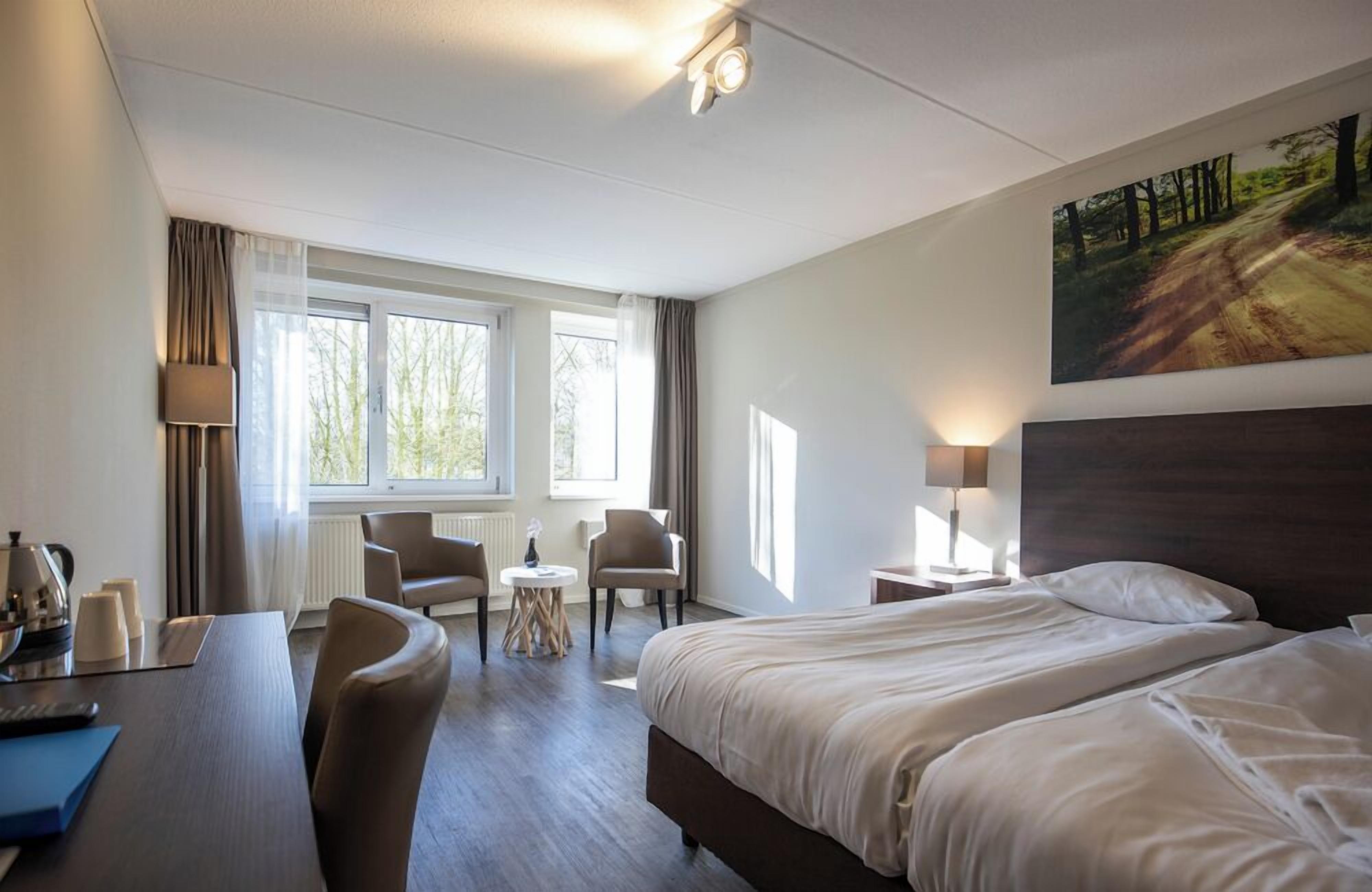 Una habitación de hotel con una cama, dos sillas, un escritorio, una lámpara y un cuadro en la pared.
