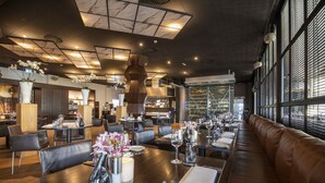 Un restaurante moderno con techos altos, conductos expuestos y una variedad de espacios para comer. Las mesas están dispuestas con manteles blancos, cubiertos y copas.