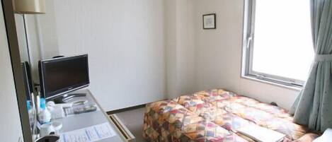 A hotel room with a bed, a desk with a computer, a window with curtains, and an air conditioning unit.