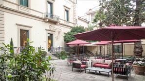 Ein Cafébereich im Freien mit Tischen und Stühlen, umgeben von einem Garten und einem Gebäude mit Balkon.