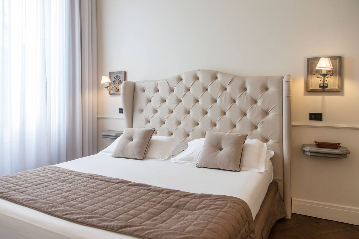 Una cama bien hecha con un cabecero capitonado, dos mesitas de noche con lámparas y una ventana con cortinas.