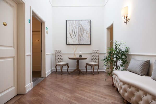 A hotel lobby with a wooden floor, a round table with two chairs, a sofa, a wall-mounted painting, and a potted plant.