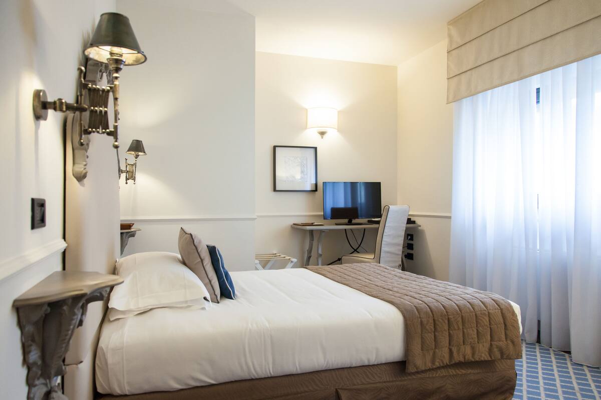 Habitación de hotel con cama, escritorio con computadora, silla, lámpara y ventana con cortinas.