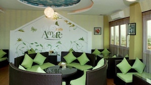 A cafe with wicker furniture and green cushions, a decorative ceiling, and framed pictures on the walls. - Amore Hotel (Hanoi)