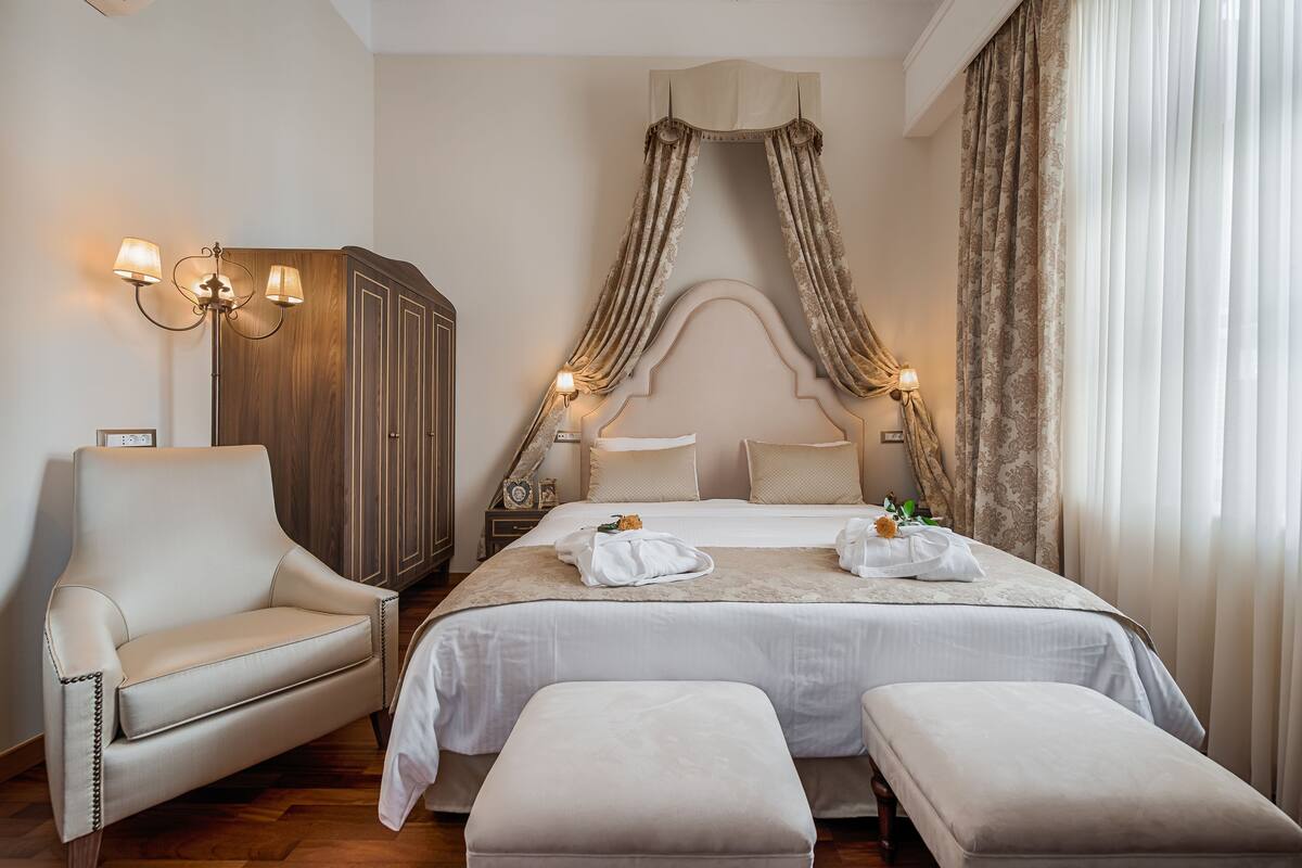 A hotel room with a large bed, a sitting area with a chair, a wardrobe, and a canopy bed with curtains.