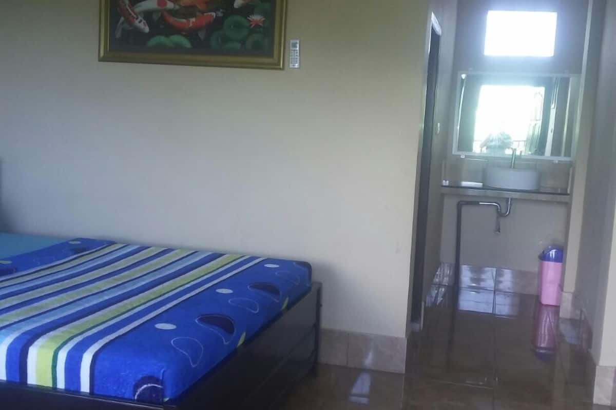 A bedroom with a bed, an air conditioning unit, a painting, a bathroom with a sink and mirror, and a tiled floor.