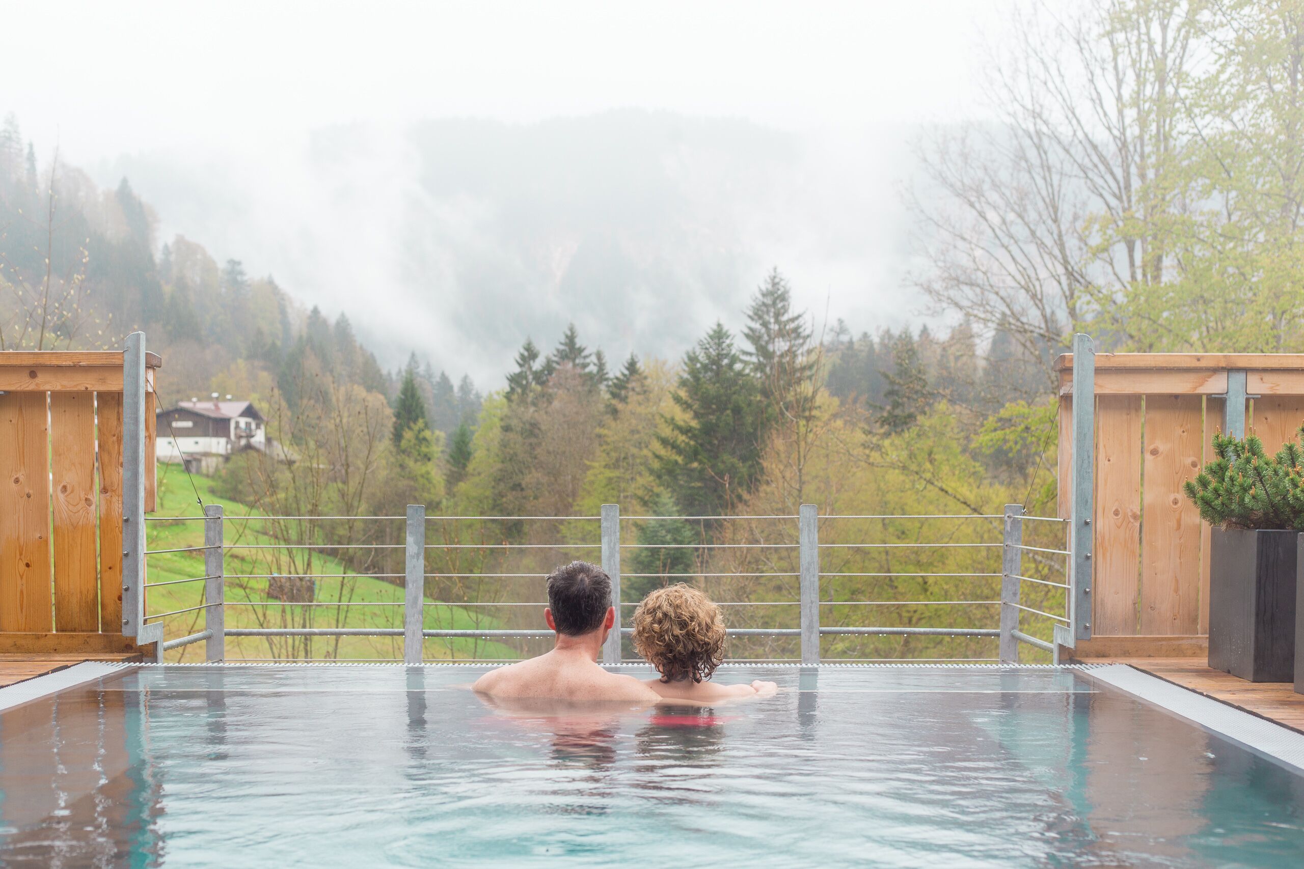 A couple in a private outdoor pool with a view of a forested hillside and a house in the background.