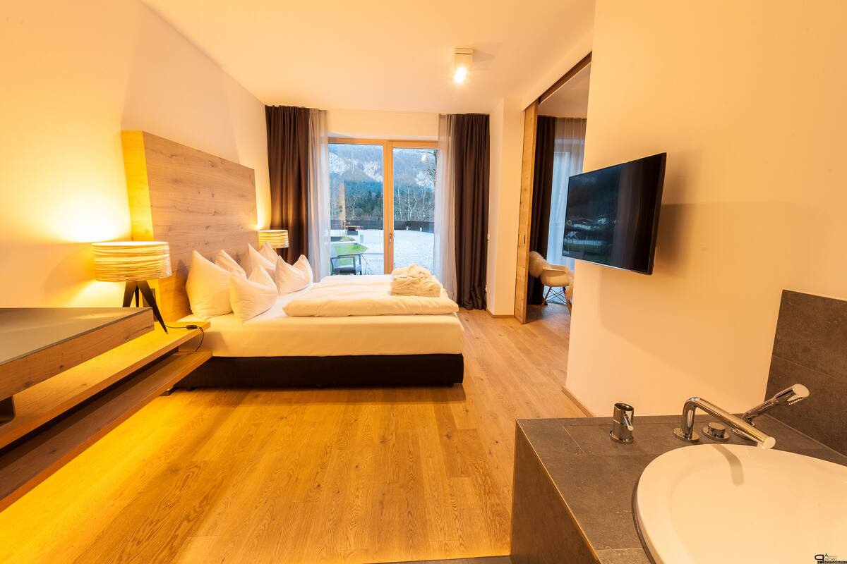 Een moderne hotelkamer met een groot bed, een flatscreen televisie, een houten hoofdbord, een nachtkastje met lampen en uitzicht op de bergen door de schuifglazen deur.
