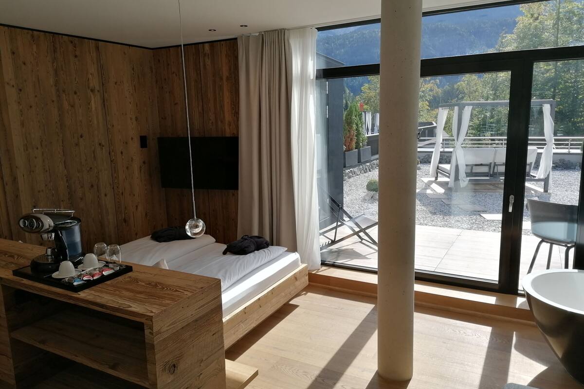 Een moderne kamer met een bed, een houten bureau, een televisie en een groot raam met uitzicht op een terras en groen.