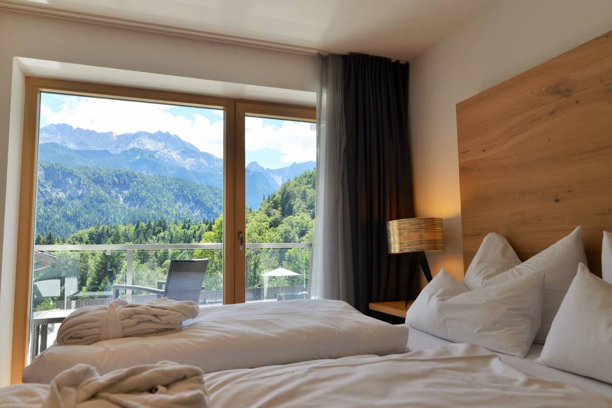 Een hotelkamer met een bed, nachtkastje, lamp en uitzicht op bergen door een groot raam.